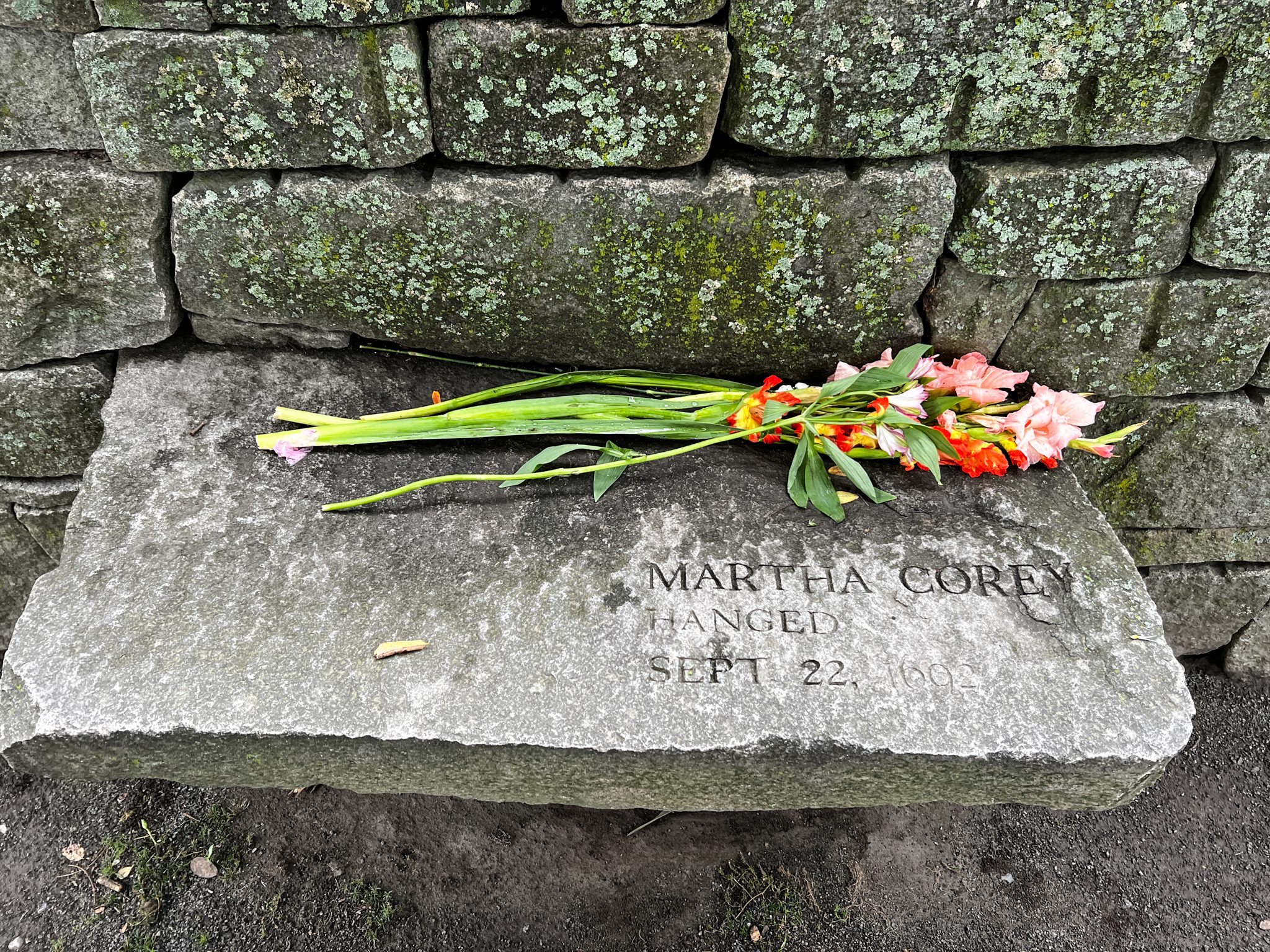 Memorial bench for Martha Corey, with a bouquet of pink and red flowers above her name, which is carved into the stone bench. 